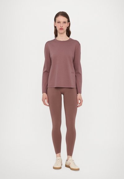 Braunes Langarmoberteil mit lockerer Passform, kombiniert mit passenden braunen Leggings. Strukturierter Stoff mit dezentem Branding auf dem Oberteil. Helle Sneakers.