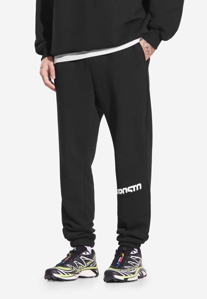 Czarne spodnie dresowe z elastycznymi ściągaczami, bocznymi kieszeniami oraz białym logo na lewej nodze. Noszone z kolorowymi, wzorzystymi butami sportowymi.