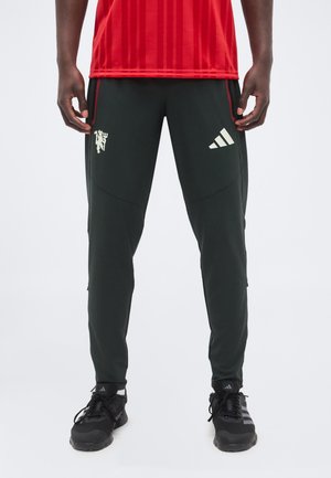 Homme portant un pantalon de sport Adidas noir avec des logos sur les cuisses, des baskets Adidas noires et un t-shirt rouge, debout devant un fond uni.
