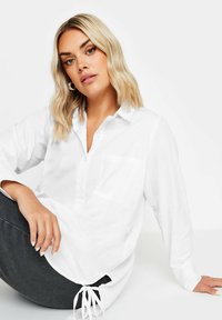 Camisa blanca de manga larga con cuello, bolsillo frontal y detalle de corbata en el dobladillo, fabricada en una tela suave. Se combina con pantalones gris oscuro.