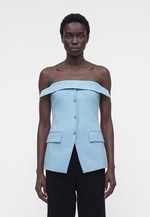 Femeie care poartă un top structurat albastru deschis, cu umerii goi, nasturi și buzunare cu clapete, asortat cu pantaloni negri cu picioare largi, privindu-se înainte.