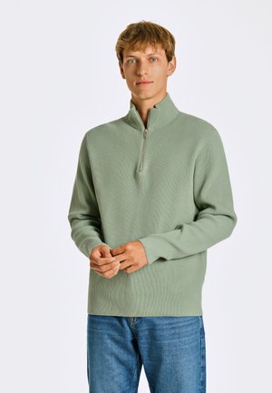 Hellgrüner Ripppullover mit halbem Reißverschlusskragen, langenÄrmeln und lockerer Passform, kombiniert mit blauen Jeans.