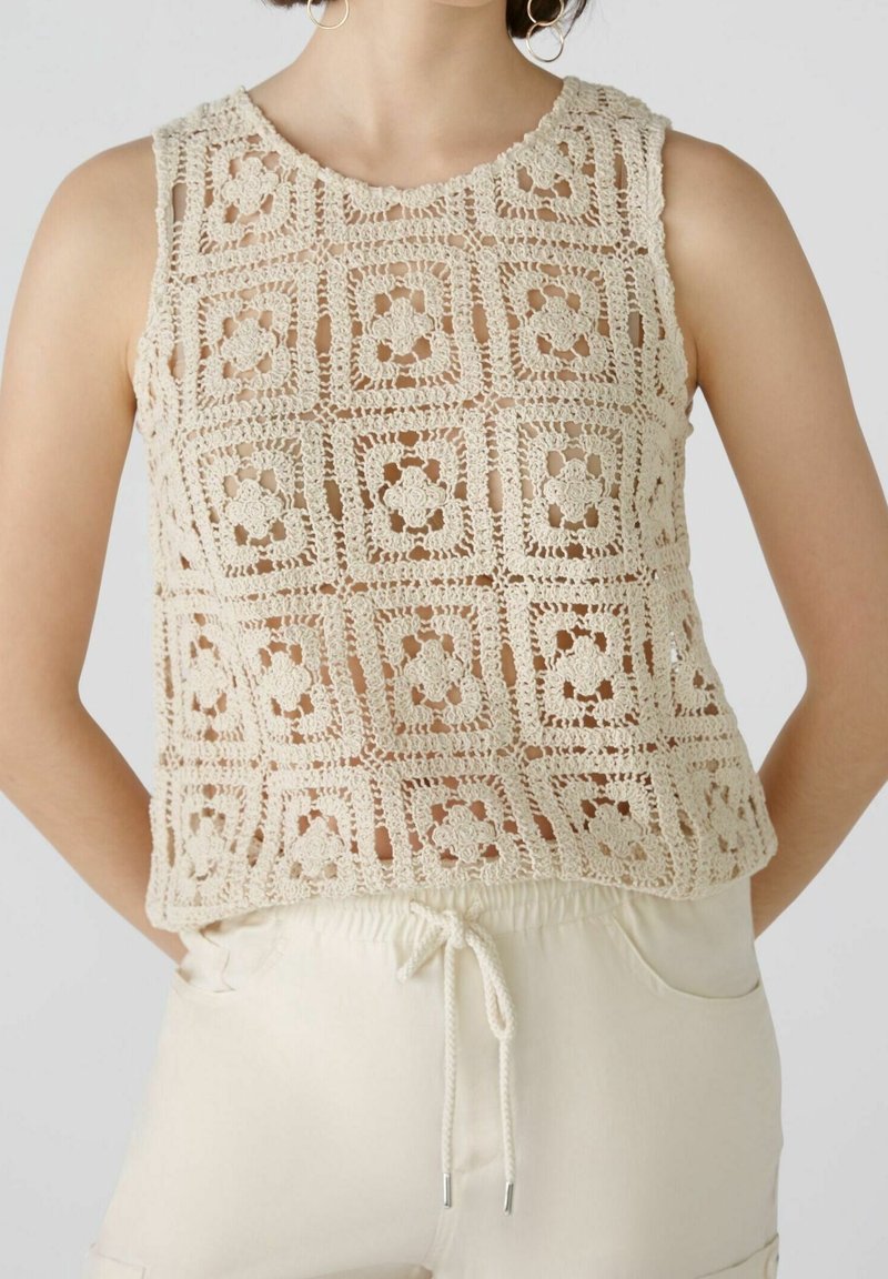 Top sans manches beige en crochet avec des motifs floraux carrés porté sur un pantalon crème clair à cordon.