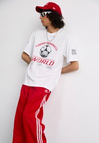 Hvit t-skjorte med rød grafikk med fotballdesign, kombinert med løse røde joggebukser med hvite striper og logo. Rød caps og solbriller.