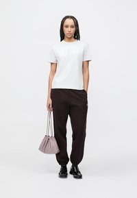 Wit katoenen T-shirt met een geperst logo, gecombineerd met donkerbruine joggingbroek. Model draagt een zacht lavendelkleurige tassen met trekkoord. Zwarte schoenen maken de look compleet.