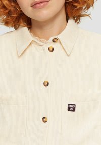 Romige corduroy overhemd met knoopsluiting, puntige kraag, twee borstzakken en een "HAPPY QS" logo patch.