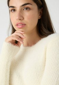 Mujer joven con cabello castaño que lleva un suéter suave y esponjoso color crema, apoyando su barbilla en la mano y mirando ligeramente hacia un lado.