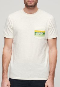 Camiseta de color crema con cuello redondo, mangas cortas y un bolsillo en el pecho que presenta un diseño de logo a rayas verdes y amarillas.