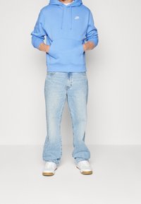 Ljusblå hoodie med framficka och vit logotyp, i kombination med ljus tvättade denimjeans och vita sneakers med beige detaljer.