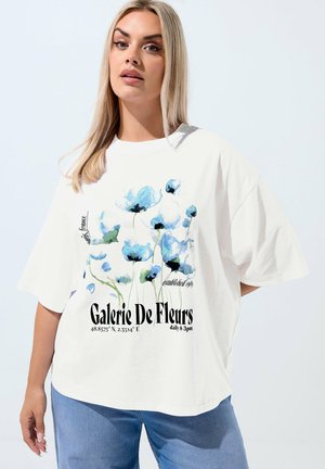 Femme blonde portant un T-shirt blanc surdimensionné avec un motif floral bleu et le texte "Galerie De Fleurs", associée à un jean bleu, sur un fond clair.