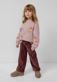 Różowy sweter z dzianiny z motywami misiów noszących czerwone czapki, zestawiony z brązowymi spodniami korkowymi. Dziecko nosi brązowe buty wsuwane.