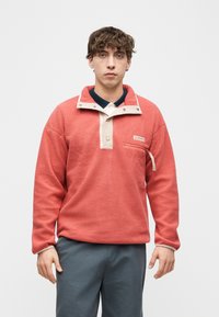 Νέος άνδρας που φοράει ένα κοραλλί φλις πουλόβερ με μπεζ κουμπιά και τσέπη στο στήθος, στέκεται μπροστά σε απλό λευκό φόντο.