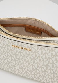 Witte stoffen tas met een MK-monogram patroon, een gouden ritssluiting en een bruine binnenvoering met een merklabel. Textuurelementen aan het oppervlak zijn zichtbaar.