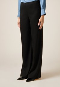 Zwarte krijtstreepbroek met een wijd-uitlopende pasvorm, gemaakt van een gladde stof; gecombineerd met een lichtblauw blouse en zwarte puntige schoenen.