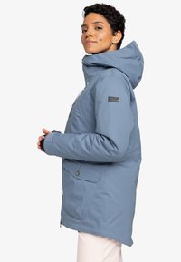 Light blue jacket with a hood, featuring a front zipper, two side pockets, and a logo patch on the sleeve. Textured fabric finish.