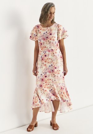 Kobieta w kremowej sukience midi w kwiaty z falbankowymi rękawami i asymetrycznym dołem, w połączeniu z brązowymi sandałami na blokowym obcasie z odkrytymi palcami.