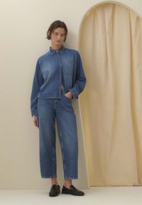 Jeanshemd mit Kragen, Brusttasche und Knopfleiste, kombiniert mit hoch taillierter, weiter Jeanshose. Schwarze flache Schuhe, neutraler Hintergrund.