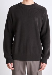 Dark brown ribbed sweater with a round neckline, textured fabric, relaxed fit, and a subtle logo patch on the chest. Soft, cozy material.