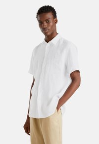 Camicia bianca in lino a maniche corte con design bottone, caratterizzata da un colletto classico e una vestibilità morbida. Indossata con pantaloni beige.