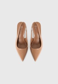 Une paire de talons aiguilles slingback en cuir verni beige à bout pointu sur un fond blanc uni, vue de dessus.