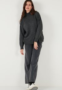 Donkergrijze gebreide trui met een hoge halslijn en geribbelde randen, gecombineerd met losse donkere jeans en witte sneakers.