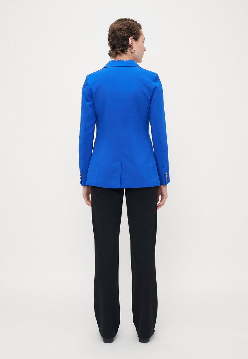 Blazer ajusté bleu avec une coupe structurée, fente simple à l'arrière, et accents de boutons dorés, associé à un pantalon noir à jambe droite.