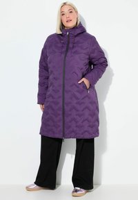 Abrigo largo acolchado morado con capucha y un patrón texturizado, cierre frontal de cremallera y bolsillos laterales, usado sobre pantalones y zapatos negros.