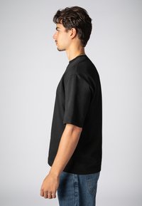 Schwarzes kurzärmliges Shirt mit rundem Halsausschnitt, aus Baumwolle gefertigt. Eine Person trägt es, kombiniert mit hellblauen Jeans, von der Seite gezeigt.
