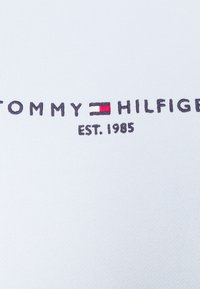 Logo Tommy Hilfiger avec drapeau rouge, blanc et bleu et texte "EST. 1985" brodé sur tissu blanc.