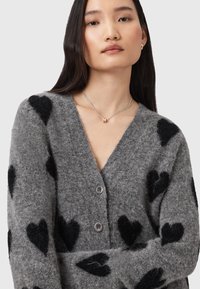 Cardigan grigio in morbido tessuto a maglia con motivi di cuori neri, scollatura a V e chiusura con bottoni. Il modello indossa una collana d'argento con pendente.