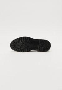Semelle de chaussure en caoutchouc noir avec un motif à clous pour une meilleure adhérence. Présente une surface texturée avec un design de bande de roulement et un renforcement autour des bords.