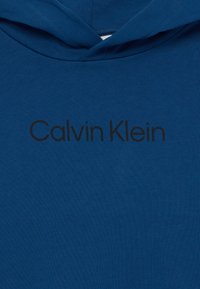 Kék pamut kapucnis pulóver, elülső kenguruzsebbel és fekete "Calvin Klein" logóval, amely a mellkas területén van nyomtatva. Sima textúra.