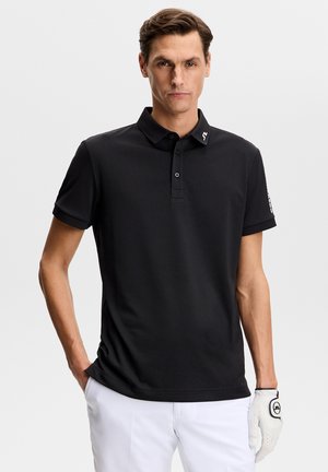 Mand iført sort kortærmet polo-shirt og hvide bukser med en hvid golfhandske på højre hånd, stående mod en ensfarvet baggrund.