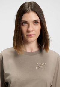 T-shirt beige à manches longues en tissu doux, avec un logo brodé subtil sur la poitrine. Encolure ronde et coupe décontractée.