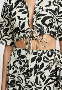 Top corto nero e crema con chiusura a coulisse, caratterizzato da un vivace motivo a foglia e maniche corte. Il design presenta dettagli a taglio.