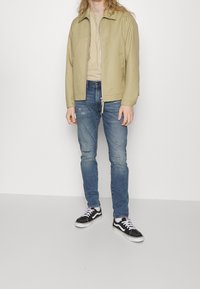 Persona llevando una chaqueta beige, una camisa beige, jeans azules desgastados, calcetines blancos y zapatillas de lona en blanco y negro, de pie contra un fondo liso.