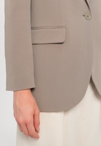 Blazer beige avec une poche à rabat et un bouton près du poignet, porté sur un pantalon blanc, montrant une main détendue sur le côté.