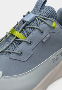 Κοντινή λήψη ανοιχτό γκρι αθλητικού παπουτσιού με επάνω μέρος από πλέγμα, κορδόνια με κίτρινη λεπτομέρεια και σύρτη, και ετικέτα "DryVent" στην πλαϊνή πλευρά.