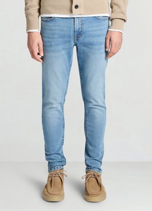 Jean en denim bleu clair à coupe slim, avec des détails délavés et un design à cinq poches, associé à des chaussures en daim couleur beige.