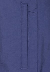 Tejido azul marino con una textura suave que presenta un bolsillo con cremallera vertical. Los bordes cosidos son visibles a lo largo de la abertura del bolsillo.