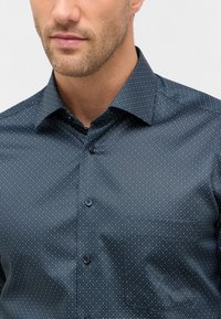 Camicia blu scuro con piccole macchie bianche e verdi. Presenta un colletto classico, una chiusura con bottoni e una singola tasca sul petto.