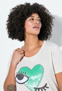 Camiseta de color crema con un diseño de corazón verde adornado con lentejuelas, complementada por un gráfico de ojo con acentos negros y pestañas.