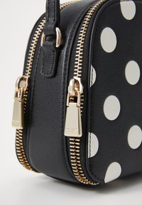 Sac en cuir noir avec des pois blancs, doté de fermetures zippées en métal doré et d'une surface texturée. Forme arrondie avec double fermeture.