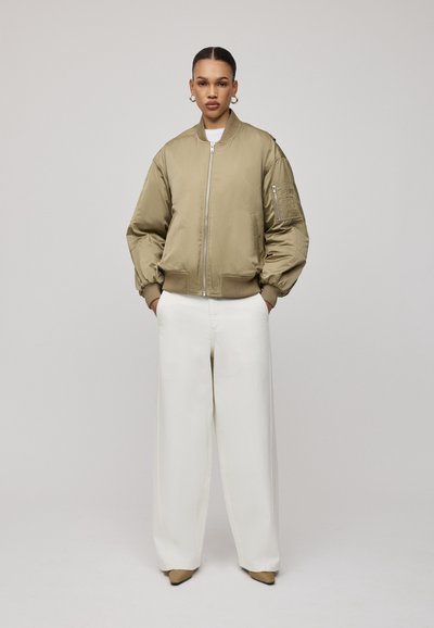 Khaki bomberka z ściągaczami na mankietach i dolnym brzegu, z zapięciem na zamek i szczegółami kieszeni po bokach, w połączeniu z szerokimi białymi spodniami i brązowymi butami.