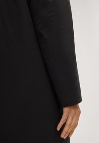 Czarny płaszcz o gładkim, matowym wykończeniu. Posiada długi rękaw o prostym kroju oraz eleganckim designie. Dłoń opiera się o bok płaszcza.