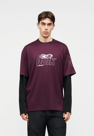 Ung mand iført en kastanjebrun T-shirt med korte ærmer og "QUIK"-logo over en sort langærmet skjorte, stående foran en ensfarvet baggrund.