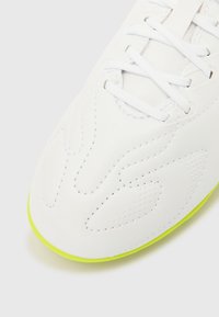 Λευκό αθλητικό παπούτσι με υφή στην επάνω επιφάνεια, με ραμμένες λεπτομέρειες και διατρήσεις. Η φωτεινή πράσινη σόλα και τα κορδόνια ολοκληρώνουν το σχέδιο.