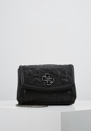 Sac à bandoulière en cuir matelassé noir avec motif embossé, logo argenté sur le rabat et chaîne argentée sur une surface blanche sur fond gris.