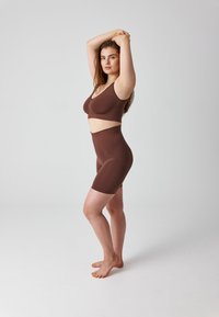 Woman in brown seamless sports bra and high-waisted biker shorts standing barefoot with arms raised and crossed above head on a white background.