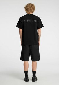 Svart t-skjorte med rund hals og grafisk tekst på baksiden, kombinert med svarte shorts. Stoffet ser glatt ut med en avslappet passform.